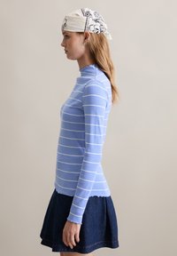 Hellblau gestreifter Rollkragenpullover aus geripptem Stoff mit weißen Streifen, kombiniert mit einem dunkelblauen, ausgestellten Jeansrock. Das Halstuch ist weiß mit schwarzem Muster.