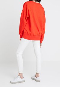 Sudadera roja de gran tamaño con hombros caídos, combinada con jeans skinny blancos y zapatillas blancas con suelas grises. Un atuendo simple y casual.
