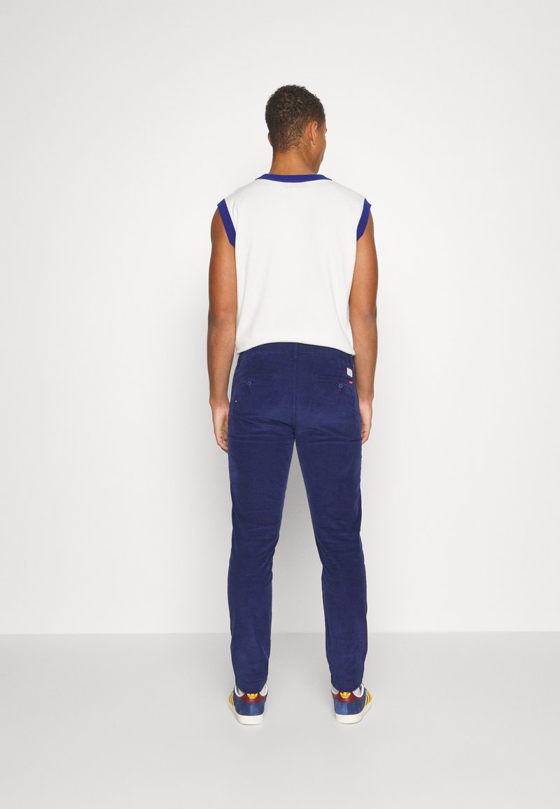 Haut sans manches ajusté blanc avec des accents bleus, associé à un pantalon en velours côtelé bleu marine. Les détails incluent des poches arrière et un tissu texturé.