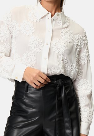 Blouse blanche boutonnée avec broderie florale texturée associée à un pantalon en cuir noir taille haute noué avec une ceinture à la taille.