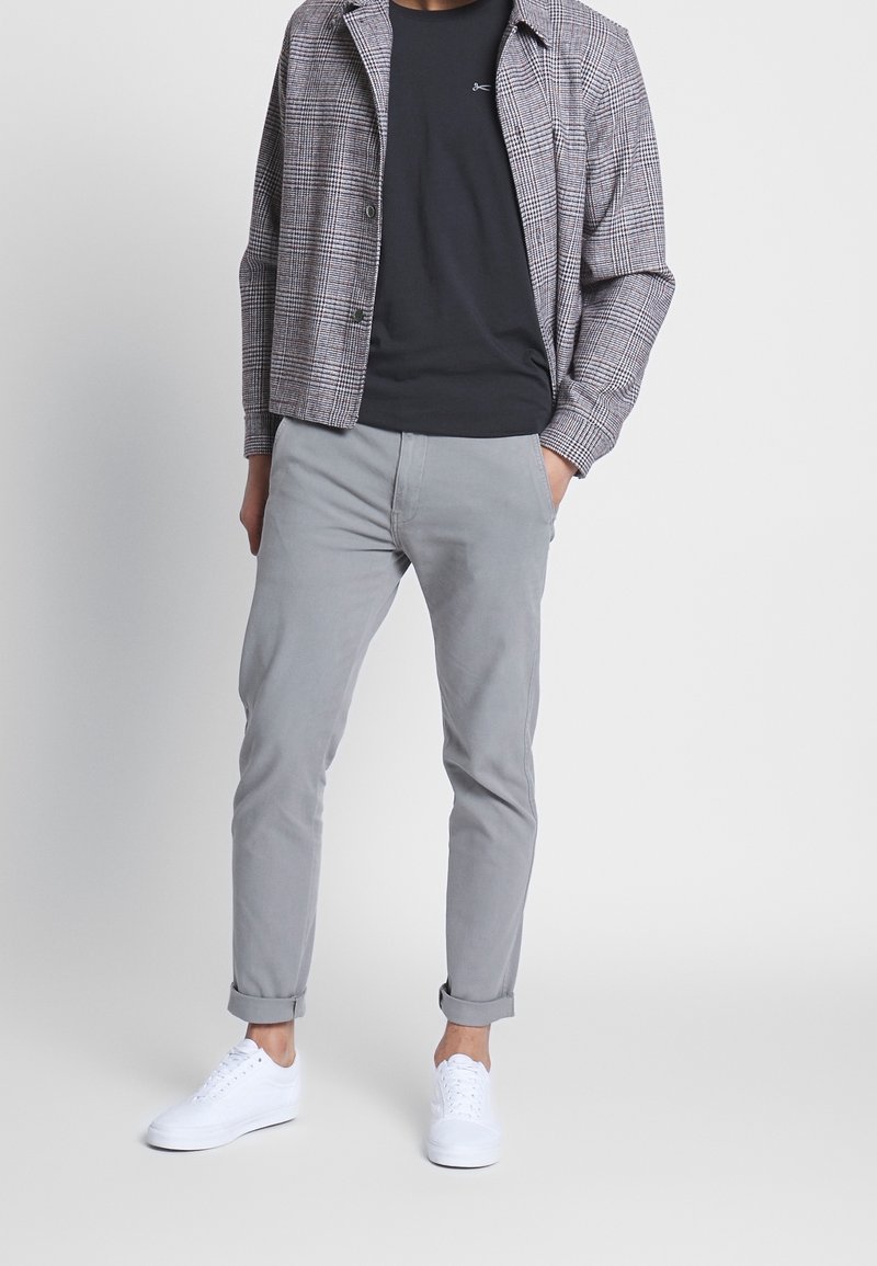 Veste à carreaux dans des tons de gris, t-shirt noir, pantalon gris et baskets blanches ; tenue décontractée avec des manches retroussées et un logo discret sur le t-shirt.
