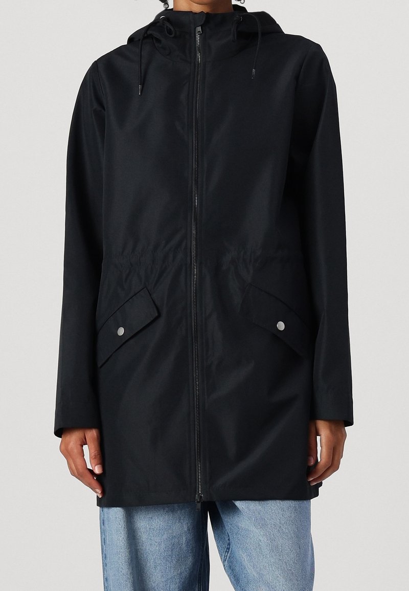 Veste de pluie noire à capuche avec fermeture éclair sur le devant et poches à rabat inclinées, portée avec un jean bleu clair.