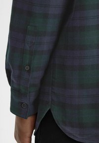 Camicia a quadri verde e blu navy, in materiale cotone, presenta polsini con bottone e un orlo curvo, mettendo in evidenza la texture e il motivo del tessuto.