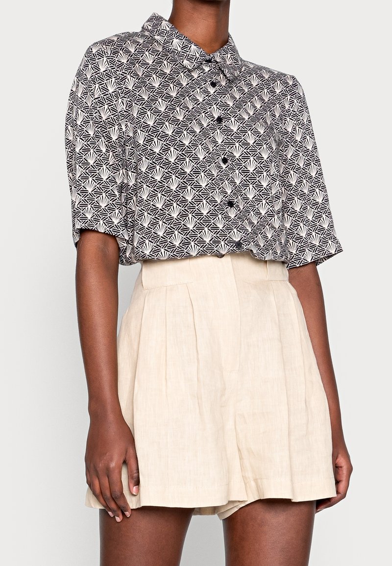 Korte ærmer knap- op skjorte i sort og hvidt geometrisk mønster, parret med højtaljede lysecreme farvede linned shorts med folder.