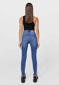 Jeans skinny en denim bleu foncé avec une taille haute, deux poches arrière et une patch en cuir beige au niveau de la taille. Des chaussures à plateforme noires sont portées.