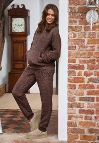 Brun fleece lounge sæt bestående af en hættebluse med kængurulomme og matchende bukser. Blød tekstur og afslappet pasform. Et par beige slippers.