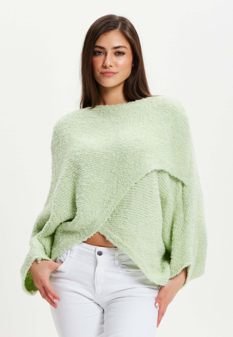 Pale green knitted sweater with a textured finish, wide sleeves, and a triangular hem, layered over white jeans.