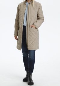 Beige vadderad lång jacka med ståkrage, framsidans dragkedja och sidofickor. Har en slät yta och diamantmönster.