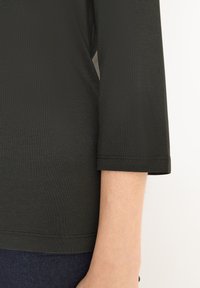 Dunkelgrünes Oberteil mit 3/4 Ärmeln, das eine glatte Textur, eine lockere Passform und einfache Saumdetails aufweist, kombiniert mit einer dunkelblauen Hose.