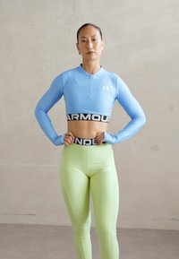Sinine pikal varrukal crop-top, millel on pool-viik ja mis on kombineeritud kõrge vöökohtadega heleda rohelise retuusiga. Must vöökoht kannab kirja "ARMOUR". Minimalistlik disain.