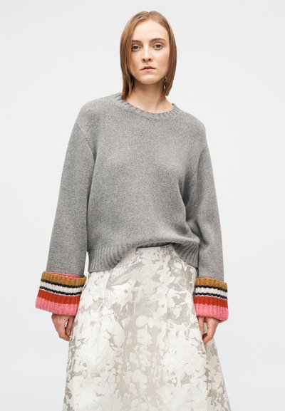 Pilkas megztas megztinis su spalvotomis dryžuotomis rankogaliais; derinamas su blizgia, gėlėto rašto, iki kelių siekiančia suknele. Paprastas, laisvo silueto dizainas.