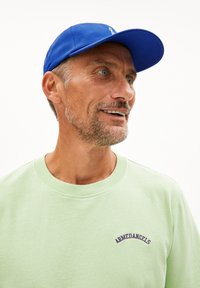 Lichtgroene sweatshirt met geribbelde halslijn en marineblauwe borduursel, gecombineerd met een blauwe pet. De pet heeft een gladde textuur en een gebogen klep.