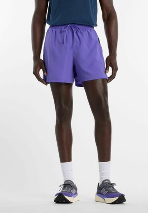 Persoon draagt paarse sportshorts, navy shirt, witte sokken en paarse en grijze hardloopschoenen, staand met het gezicht naar voren.
