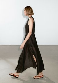 Black linen maxi dress with side slits, flat red sandals, and a loose fit. Smooth texture and minimalistic design, seen walking on a flat surface.