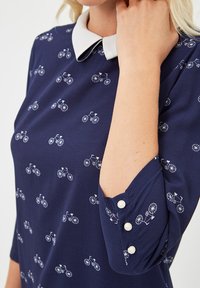 Blusa azul marino con patrones de bicicletas blancas, cuello puntiagudo blanco, tres botones de perla en el puño de la manga, llevada por una persona con la mano cerca del cuello.