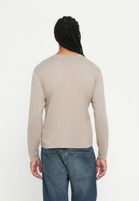 Persoon met lang gevlochten haar, gekleed in een beige shirt met lange mouwen en blauwe spijkerbroek, met de rug naar de camera tegen een eenvoudige witte achtergrond.