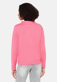 Femme aux cheveux bouclés portant un pull rose à manches longues et un jean gris clair, debout de dos sur un fond uni.