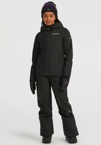 Woman wearing black winter jacket, pants, gloves, beanie, and ski goggles, standing against a plain light background.