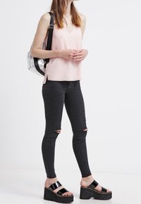 Femme portant un haut sans manches rose clair, un jean skinny noir déchiré, des sandales à plateforme noires et portant un sac à dos argenté sur une épaule.