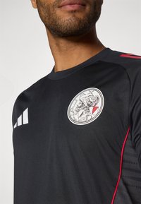 Sort atletisk skjorte med rund hals, udstyret med et fremtræden cirkulært logo, hvide detaljer og røde striber langs siderne. Glat tekstur.