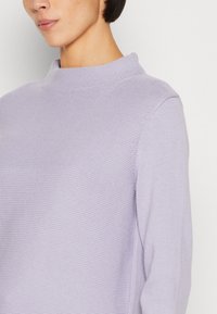 Femme portant un pull en maille texturée violet clair avec un col montant court et relevé sur un fond blanc uni.