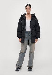 Svart dunjakke med hette, to store frontlommer og glidelås. Båret over en hvit topp og kombinert med falmede jeans.