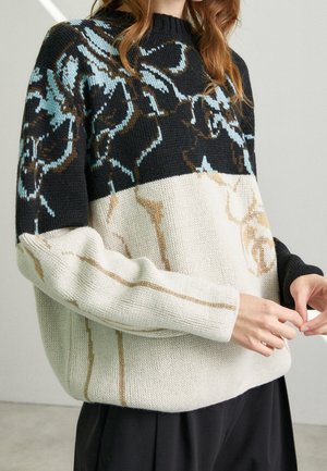 Gestrickter Pullover mit einem schwarzen Oberteil, das blaue und braune Blumenmuster zeigt, und einem cremefarbenen Unterteil mit schmalen braunen Streifen.