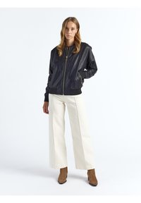 Zwarte leren bomberjack met een rits aan de voorkant, geribde manchetten en zakken, gecombineerd met witte broek met wijde pijpen en bruine enkellaarsjes.