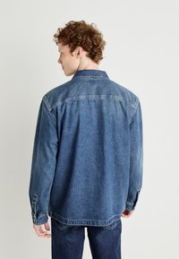 Giacca di denim di blu medio, con colletto, polsini con bottoni e vestibilità rilassata. La vista posteriore mostra i dettagli delle cuciture e una texture leggermente sbiadita.