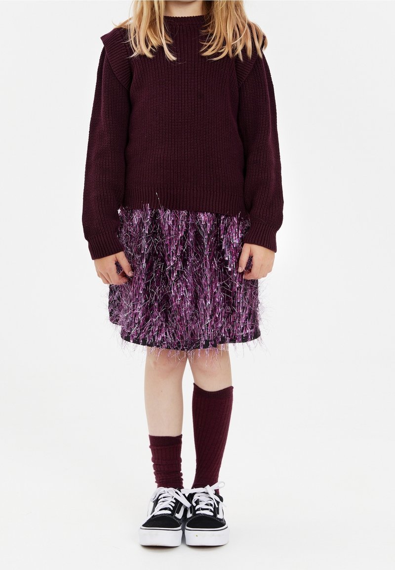 Enfant portant un pull violet foncé, une jupe violette et argentée scintillante, des chaussettes hautes dépareillées, et des baskets noires et blanches, debout devant un fond blanc.