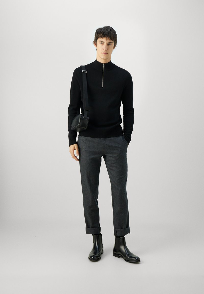 Black zip-up sweater, gray trousers with rolled cuffs, and black ankle boots. Model carries a small black shoulder bag. Plain background.