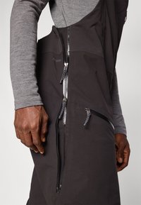 Juodos vandeniui atsparios kombinezonas su keliais užtrauktukais, matinio paviršiaus ir tekstūruoto audinio, dėvimas su pilku ilgu rankovėmis marškinėliu po apačia.