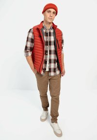 Red puffer vest over a plaid shirt, tan pants, and white sneakers. The vest features a black zipper and ribbed knit beanie.