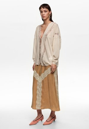 Joven mujer con cárdigan de encaje color crema sobre falda midi color coñac con detalles de encaje y zapatos bajos de tacón coral puntiagudos, de pie contra un fondo blanco.