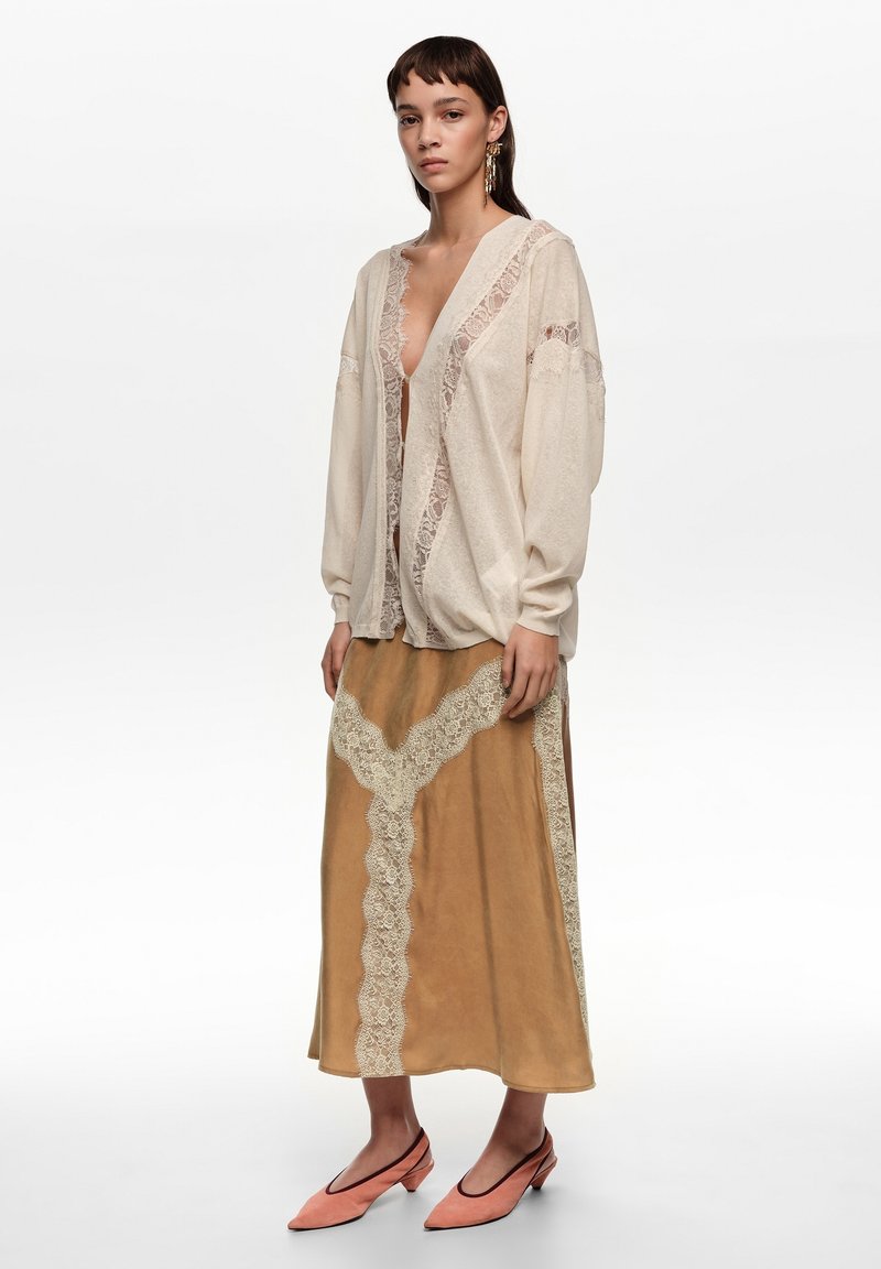 Jeune femme portant un cardigan en dentelle crème sur une jupe midi beige avec des détails en dentelle et des chaussures corail à talons bas et pointues, debout devant un fond blanc.