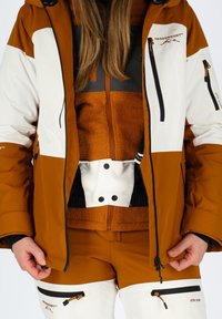 Giacca da sci marrone e bianca con fodera interna in pile arancione a trama e accenti neri; i pantaloni presentano tessuto bianco con tasche con cerniera.