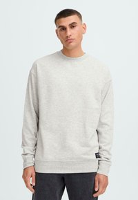 Bărbat cu păr scurt, purtând un pulover gri deschis cu guler tip crewneck și pantaloni închiși la culoare, stand pe un fundal alb simplu.