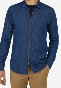 Camicia in denim blu con maniche lunghe, frontale con zip, colletto a punta e una superficie liscia. Si vede un abbinamento con pantaloni beige.