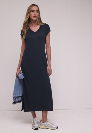 Femme en robe longue bleu marine col en V et baskets argentées tenant une veste en jean bleu clair pliée sur fond uni.