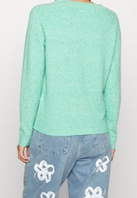 Mintgrüner Strickpullover mit lockerem Schnitt und geripptem Saum, kombiniert mit hellblauen Jeanshosen mit weißen Blumenmustern.