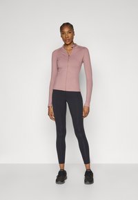 Light pink long-sleeve zip-up athletic top paired with black leggings and black sneakers. Features a fitted design and smooth texture.