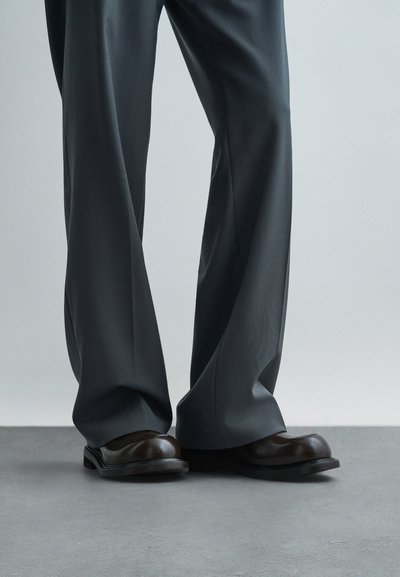 Gambe che indossano pantaloni larghi grigio scuro e scarpe di pelle nera lucida, in piedi su un pavimento grigio liscio contro uno sfondo neutro.