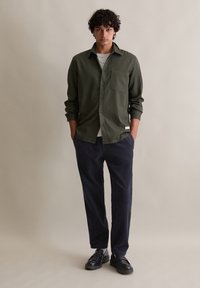 Giovane uomo in piedi con le mani in tasca, indossa una camicia verde oliva con bottoni, una maglietta bianca sotto, pantaloni di velluto a coste blu scuro e scarpe di pelle nera.