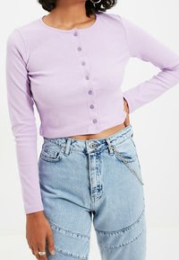 Lila pitkähihaine croptop, jossa on pyöreä kaula-aukko ja kuusi nappia edessä, yhdistettynä korkeavyötäröisiin vaaleansinisiin farkkuihin, joissa on ketjuyksityiskohta.