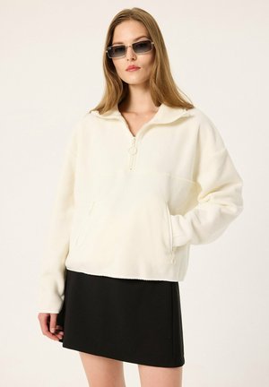 Femme portant des lunettes de soleil rectangulaires, un pull en polaire blanc avec un col zippé et une jupe noire, debout sur un fond uni.
