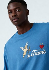 T-shirt en coton bleu avec un chérubin ailé brodé, tenant un arc. Le texte indique « je t’aime » en écriture blanche avec un accent de cœur rouge.