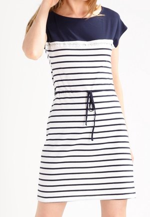 Kobieta ubrana w granatowo-białą sukienkę w paski sięgającą do kolan, z krótkimi rękawami i wiązaniem w pasie.
