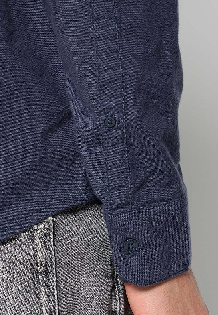 Navyblaues Hemd mit Knopfleiste und gewebter Textur, ausgestattet mit einem geknöpften Armabschluss und einem dezenten Markenetikett. Der Stoff wirkt strapazierfähig und glatt.