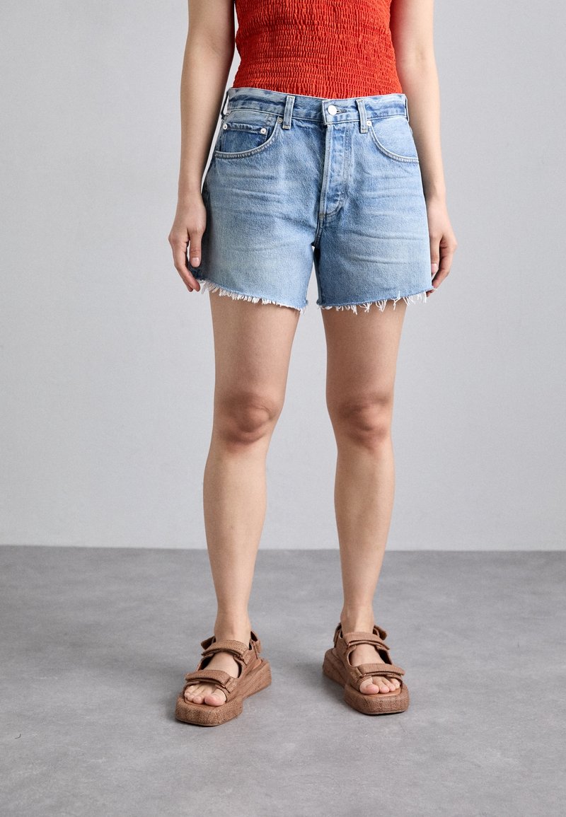 Woman wearing light blue denim shorts, red sleeveless top, and brown open-toe sandals standing on gray floor against a gray wall.