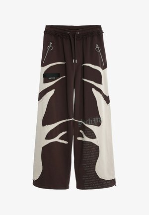 Pantalones de pierna ancha en marrón oscuro y crema con patrones de árboles abstractos, bolsillos con cremallera y cintura ajustable con cordón. Detalles de texto en la parte inferior de la pierna.